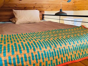 A bedroom with a wooden ceiling, a bed with a patterned blanket, a pillow, a bedside lamp, and a laundry basket.