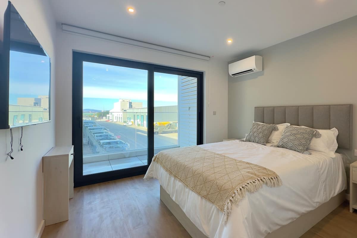 A bedroom with a large bed, a nightstand, a TV, and a view of a street and buildings through a sliding door.