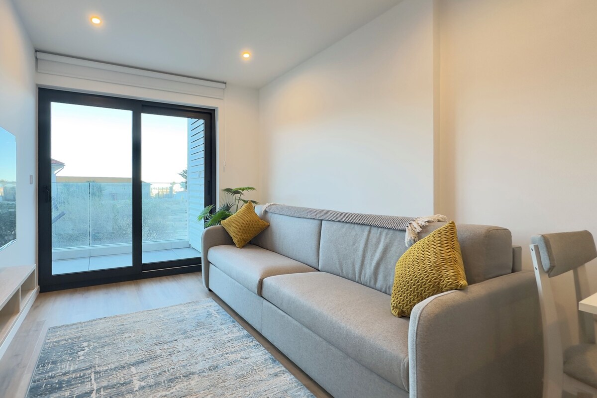A modern living room with a sofa, a flat-screen TV, and a sliding glass door leading to a balcony.