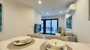 A modern living room with a dining area, a sofa, a TV, and a large window. - Victory Suites (Gibraltar)