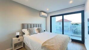 A bedroom with a bed, bedside table, lamp, air conditioning unit, and a large window with a view of buildings. - Victory Suites (Gibraltar)