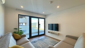A modern living room with a sofa, a flat-screen TV, and a sliding glass door leading to an outdoor area. - Victory Suites (Gibraltar)
