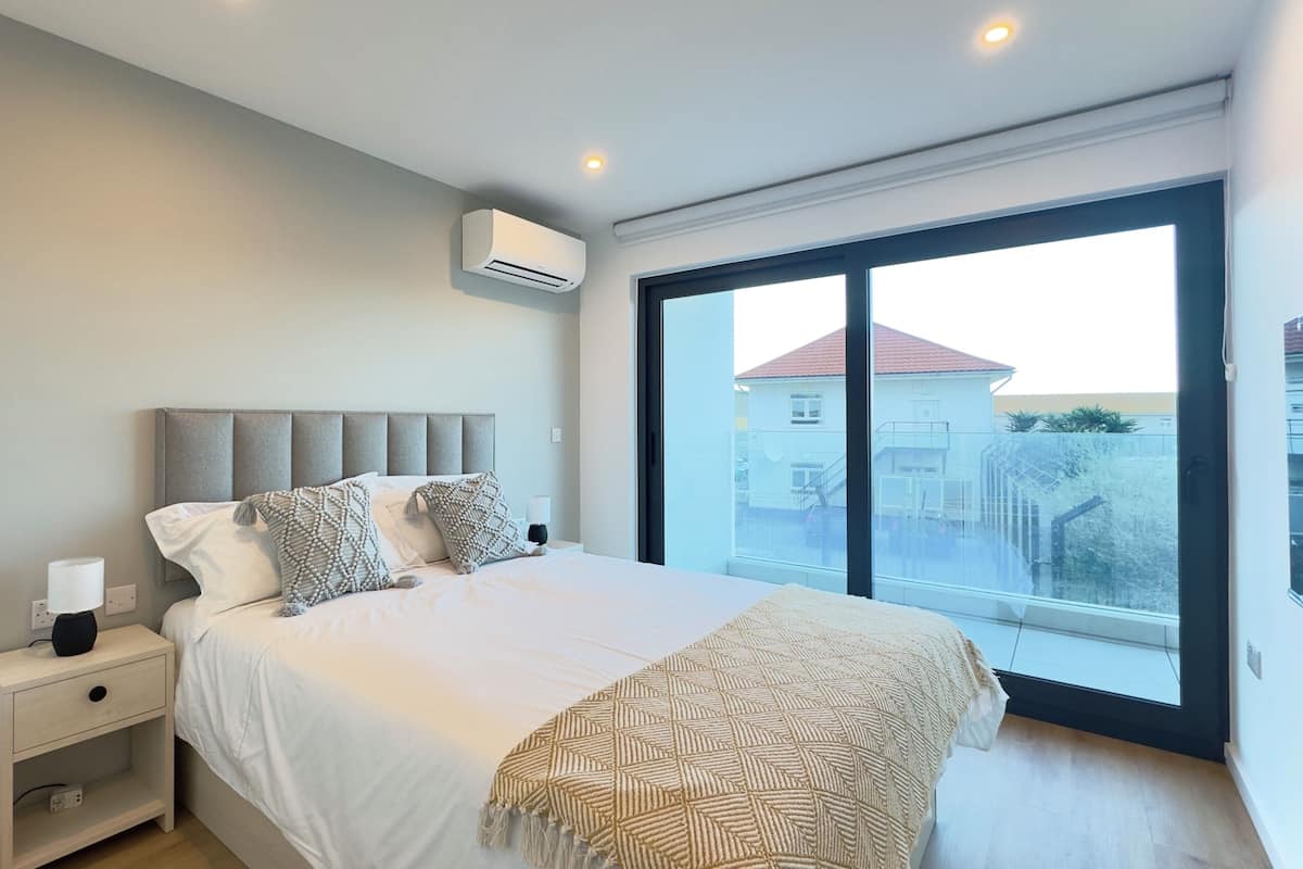 A bedroom with a bed, bedside table, lamp, air conditioning unit, and a large window with a view of buildings.