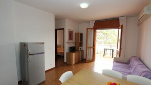 Una camera compatta con un frigorifero, un tavolo da pranzo con un cesto di frutta, un divano e vista su un'area esterna tramite una porta.