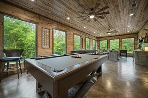 Mesa de billar en una habitación con paredes de madera y ventanas amplias, un ventilador de techo y una zona de bar.