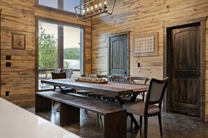 Un interior rústico de madera con una gran mesa de comedor, bancos y sillas, una araña y vistas a través de amplios ventanales.