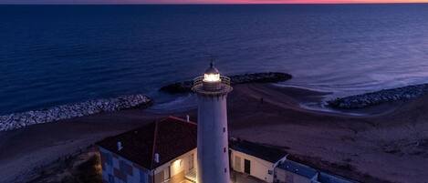 A lighthouse with a circular base and a dome, situated on a beach with a rocky breakwater.