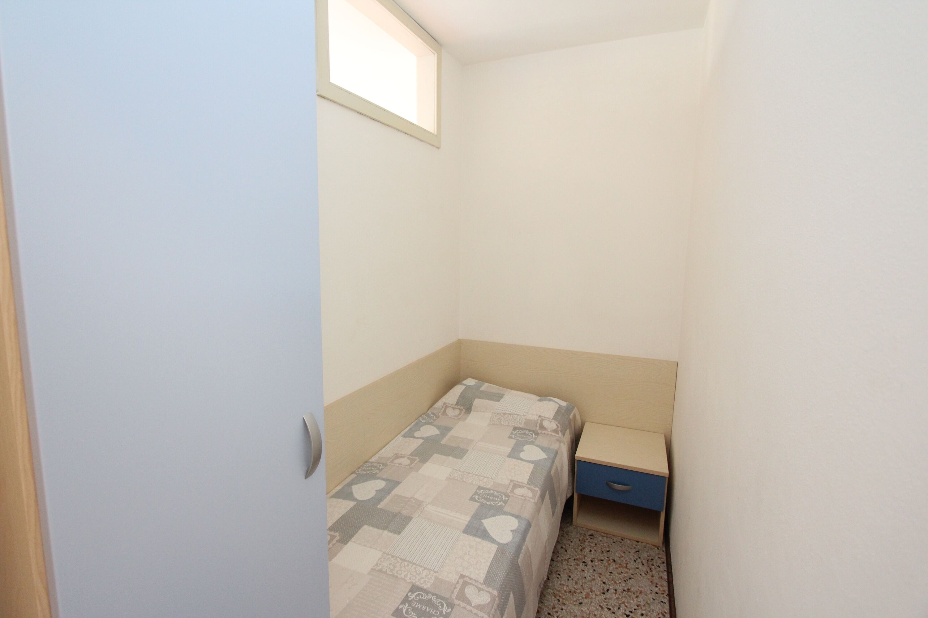 A small, narrow room with a single bed, a nightstand, and a small drawer.