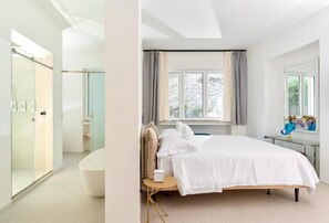 Une chambre moderne avec un grand lit, une table de chevet, une chaise, une fenêtre avec des rideaux et une salle de bain dotée d’une cabine de douche en verre.