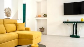 Un salon moderne avec un canapé d’angle jaune, une table basse ronde noire, une console verte agrémentée d’objets décoratifs et un téléviseur à écran plat fixé au mur.