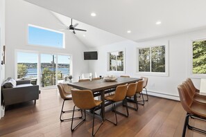 Une salle à manger spacieuse avec une grande table et six chaises, un ventilateur de plafond et de grandes fenêtres offrant une vue sur un lac et des arbres.