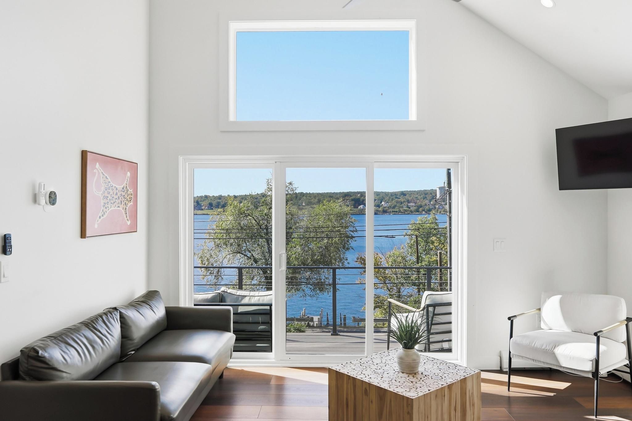 A modern living room with a leather sofa, a white armchair, and a coffee table. A large window offers a view of a lake and trees.