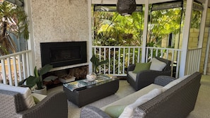 A covered outdoor seating area with wicker furniture, a flat-screen TV, and a view of lush greenery.