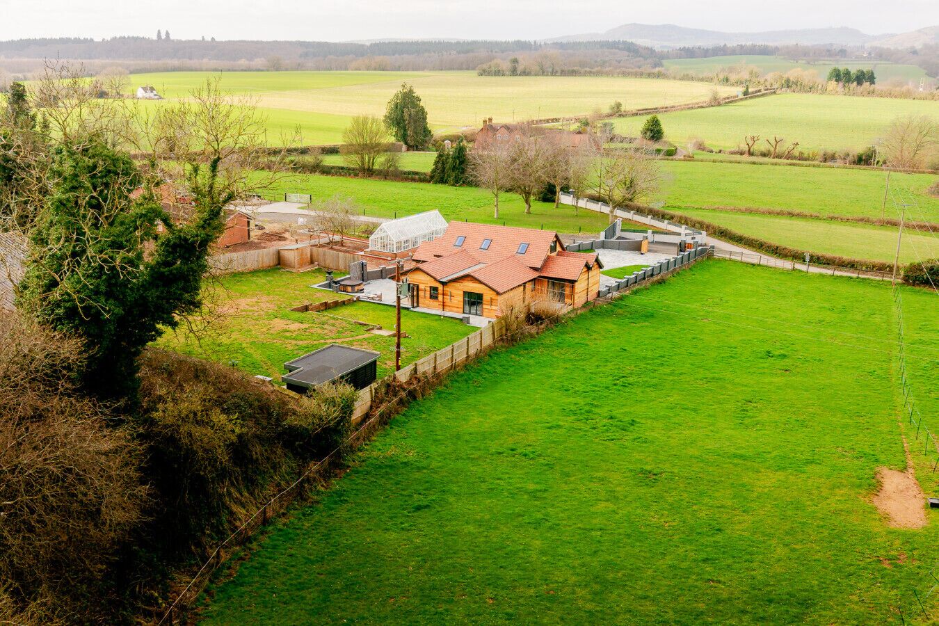 A rural house with a fenced yard, a greenhouse, and a large open field in the background.