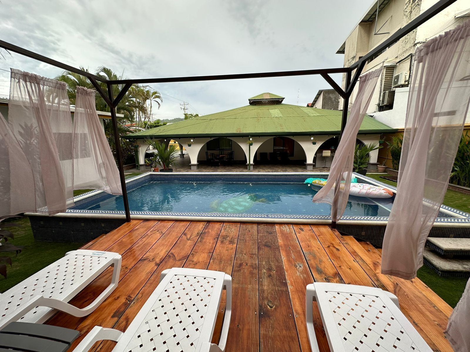 A pool area with a wooden deck, white lounge chairs, and a covered patio with arches.