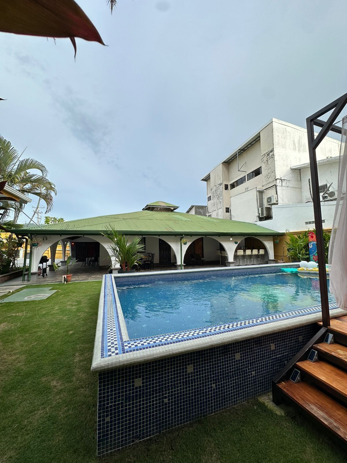 Une piscine avec un bord carrelé, un patio couvert et un bâtiment en arrière-plan.