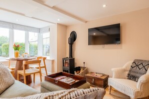 A living room with a fireplace, a television, a dining area with chairs, and a view through large windows. - The Cowshed | Banham Cottages (Bourton-on-the-Water)