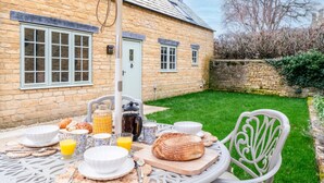 A breakfast setup with croissants, bread, and juice on an outdoor table, with a stone house and a grassy yard in the background. - The Haybarn | Banham Cottages (Bourton-on-the-Water)