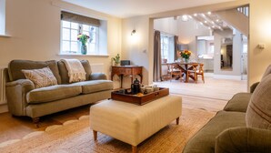 A living room with a sofa, ottoman, and a dining area with a table and chairs. - The Haybarn | Banham Cottages (Bourton-on-the-Water)