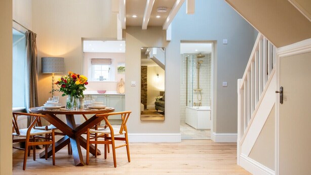 A dining area with a round table and chairs, a vase with flowers, and a staircase leading to another level. - The Haybarn | Banham Cottages (Bourton-on-the-Water)