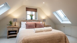 Ein Schlafzimmer mit schrägen Wänden, einem Oberlicht, einem Bett mit gemusterten Kissen, Nachttischen mit Lampen und einem Fenster mit Blick.