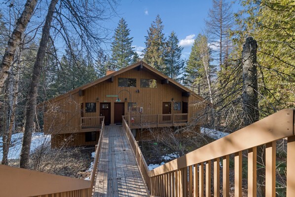A wooden cabin with a deck leading to the entrance, surrounded by trees. - Ptarmigan Village | Lake and Hot Tub Access (Whitefish)