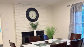 A dining area with a brick wall, a round clock, a fireplace, and a window with curtains. - Cozy, Charming and very Convenient House (Spartanburg)