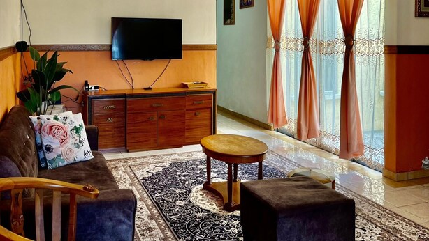 Un salon doté d’un ventilateur de plafond, d’un téléviseur à écran plat, d’un canapé, d’une table basse, d’une table d’appoint, d’un tapis à motifs et d’une fenêtre avec des rideaux.