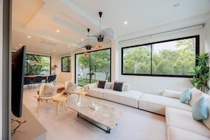 Una sala moderna con un sofá modular, una mesita de centro y un televisor. Hay ventanas amplias con vistas a zonas verdes.