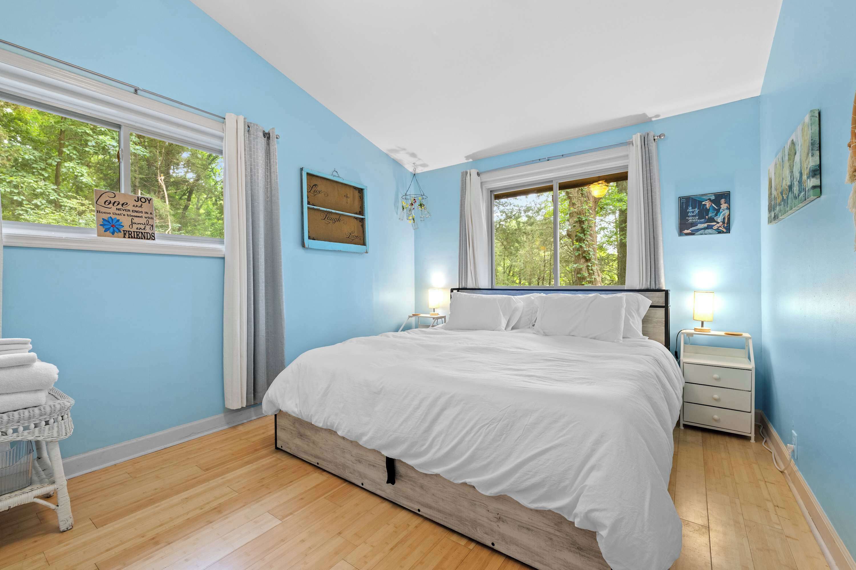 A bedroom with a bed, two bedside tables, a nightstand, a window with curtains, and a wall with a sign and a poster.