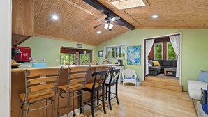 A kitchen with a wooden ceiling, a bar area with stools, a dining table, and a window with a view of trees.