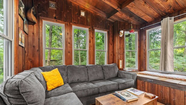 A cozy cabin interior with a gray sectional sofa, a wooden table, and large windows offering a view of the forest.