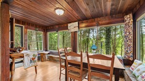 A wooden interior with a dining table and chairs, large windows, and a view of trees.
