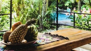 Un décor tropical avec une table en bois, un saladier rempli de fruits et une vue sur une piscine à travers une fenêtre en verre.