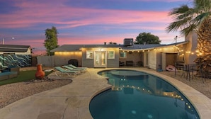 Piscina com borda curva, área de varanda coberta com espreguiçadeiras e uma casa com luzes de corda.