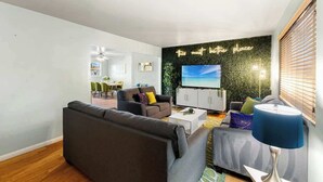 Uma sala de estar moderna com um sofá, uma mesa de centro e uma TV. Ao fundo, há uma área de jantar com cadeiras e uma mesa.
