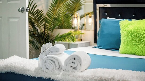 Cama bem arrumada com toalhas enroladas, cabeceira azul e travesseiro verde.