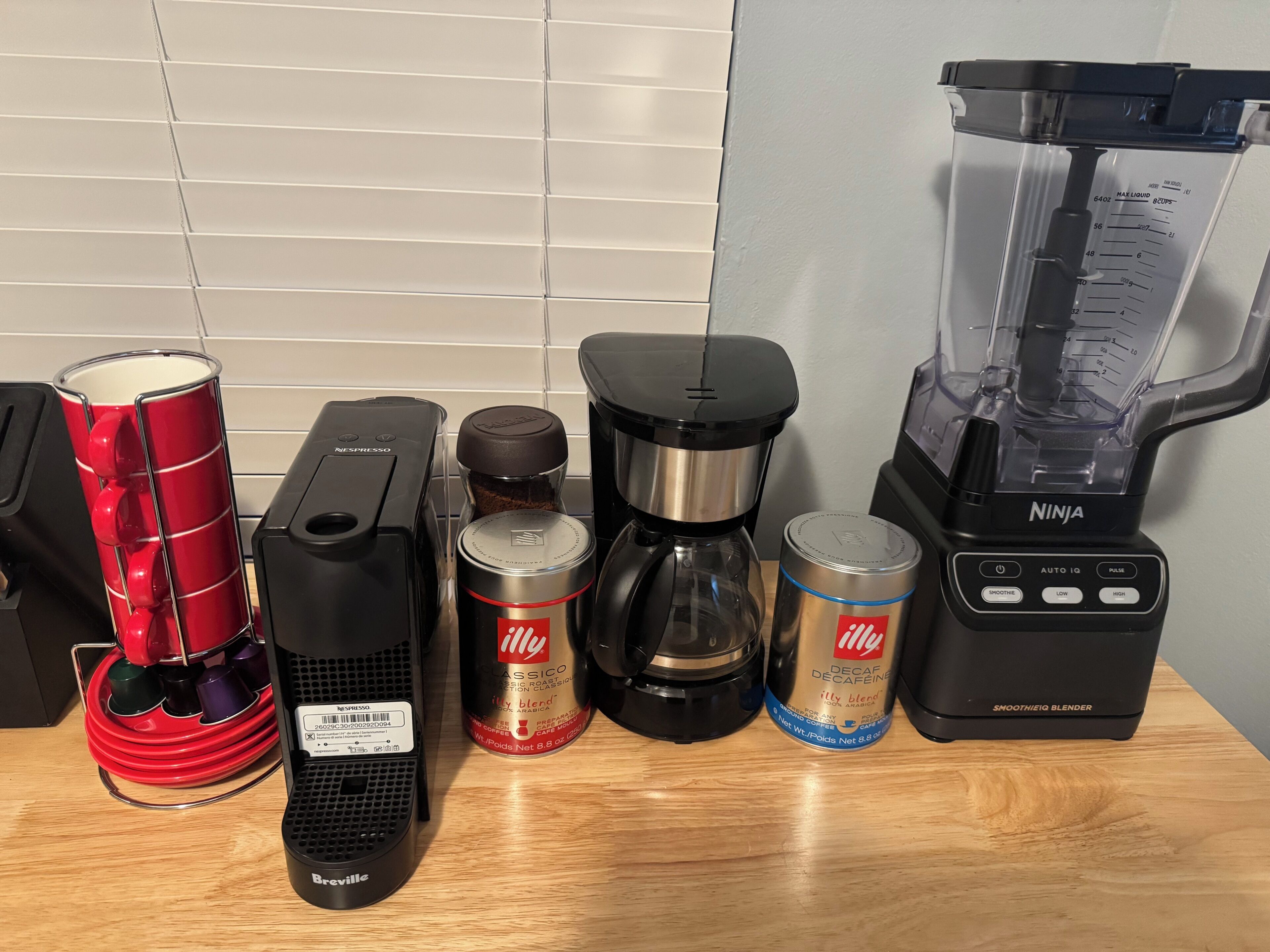 Un plan de travail de cuisine avec une machine à café, un mixeur, une cafetière, un moulin à café et une tasse rouge.