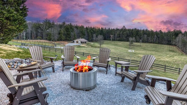Ein Lagerfeuer, umgeben von Adirondack-Stühlen, einer Schotterterrasse und Blick auf ein Wiesenfeld mit Bäumen.