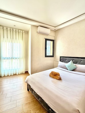 A hotel room with a bed, a window with curtains, an air conditioning unit, and a view of the outside.