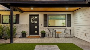 Een modern huis met een zwarte deur, twee ramen met luiken, een overdekt terras met twee stoelen en een kleine tuin.