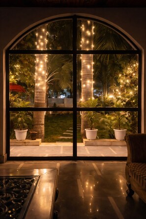 Una habitación con un gran ventanal de arco que da a un jardín por la noche, con luces colgantes y plantas en macetas.