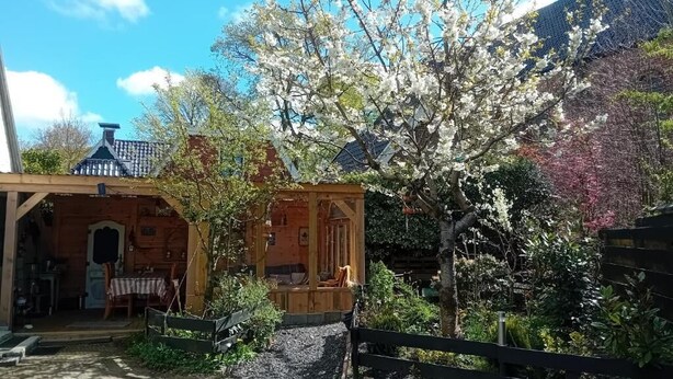 A wooden cabin with a porch, surrounded by a garden with blooming trees. - B&B De Zwerver (Oldehove)