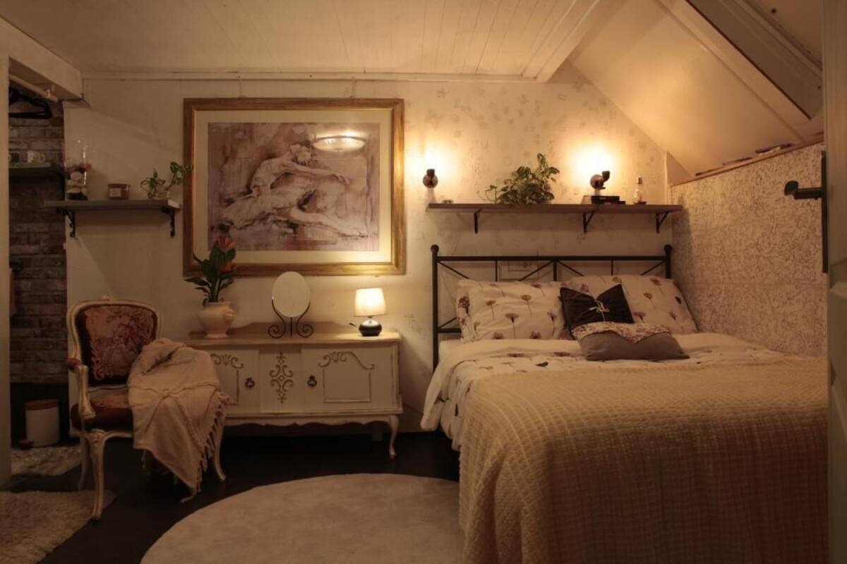 Une chambre à coucher avec un lit, une table de chevet avec une lampe, une chaise, une commode, un tableau et des étagères avec des plantes.