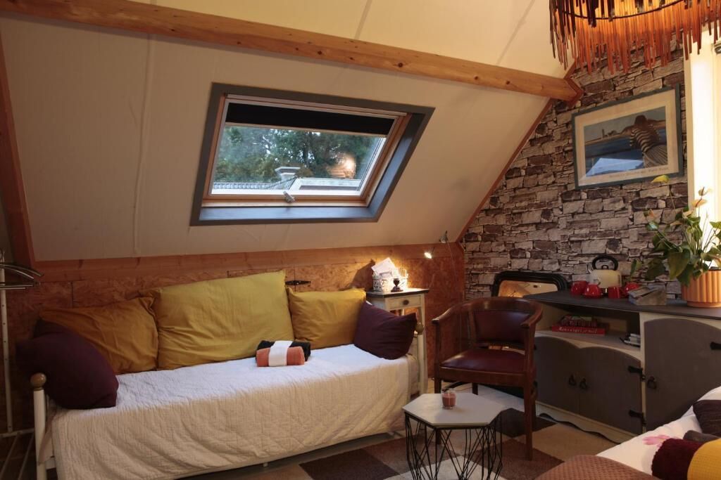 A cozy room with a sloped ceiling, a bed with pillows, a chair, a small table, and a stone wall with a picture.