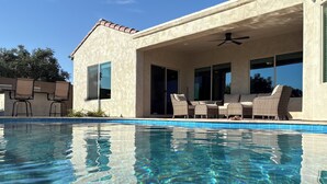 Una piscina con agua cristalina, un porche cubierto con mobiliario de mimbre y un edificio con fachada de estuco.