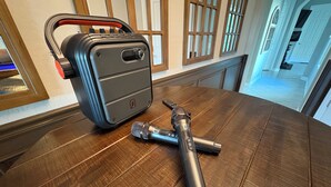 Una maleta, un micrófono y un adaptador de corriente sobre una mesa de madera.