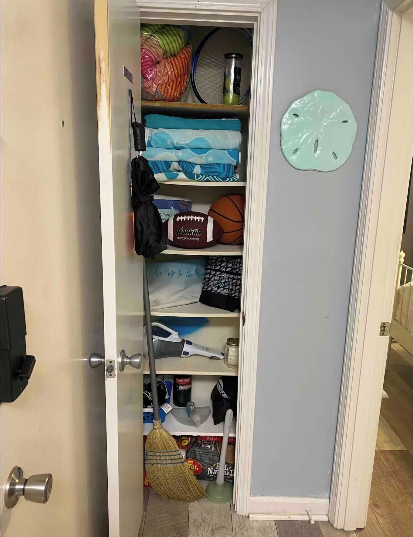 A storage closet with shelves containing folded clothes, a football, a basketball, a broom, and cleaning supplies.