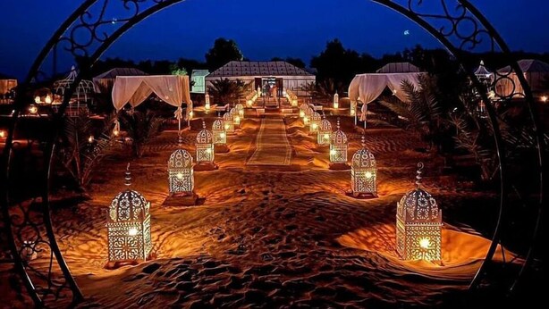 A desert pathway leading to a building with a covered entrance, surrounded by lanterns and palm trees. - Sahari Merzouga Camp (Merzouga)