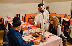A dining area with tables set for a meal, a waiter attending to guests, and a tented structure in the background. - Sahari Merzouga Camp (Merzouga)